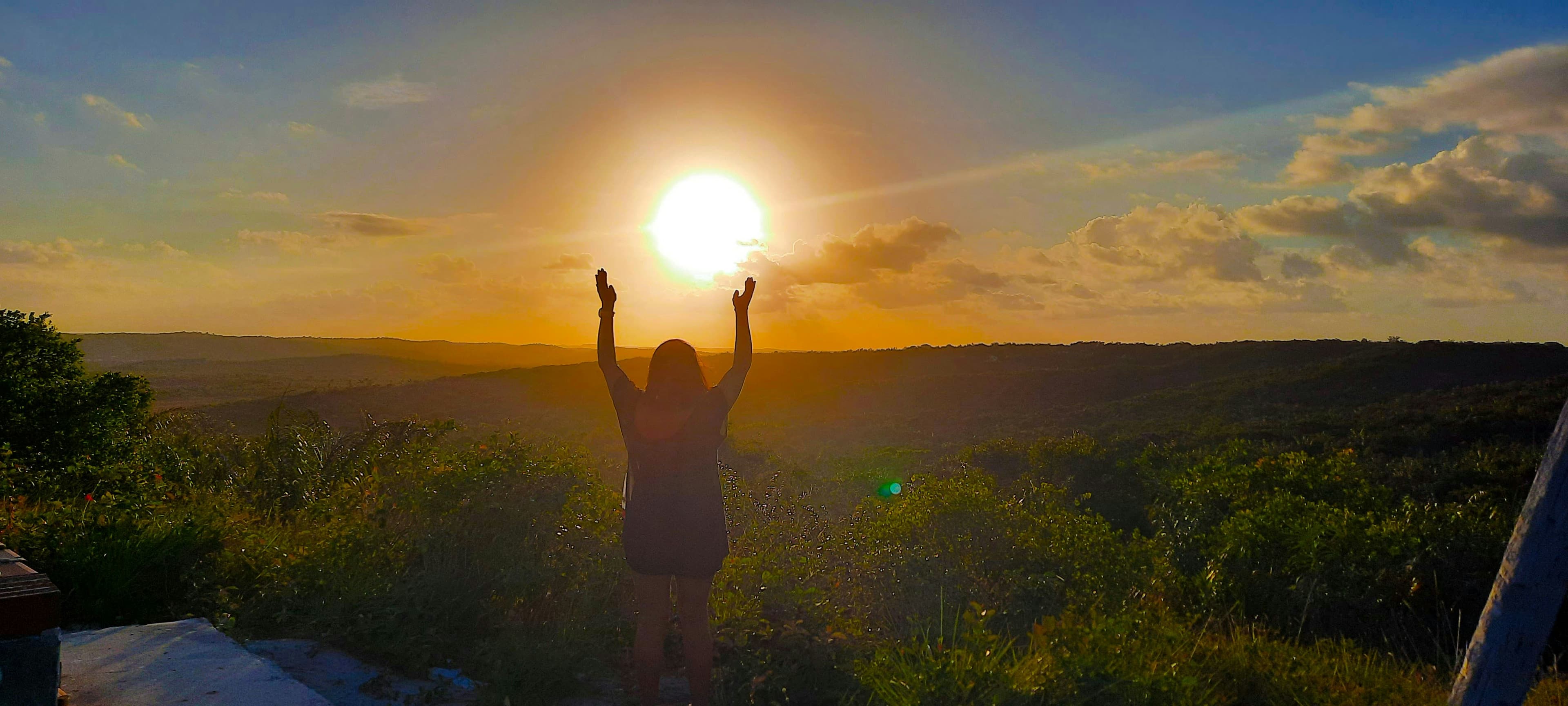 A person on a hilltop lifting their arms to a golden sunset over green hills